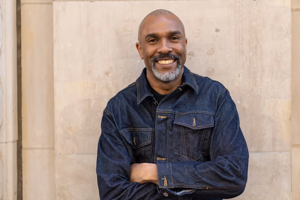 Smiling middle-aged man with gray beard wearing a dark denim jacket standing against a beige wall with arms crossed.