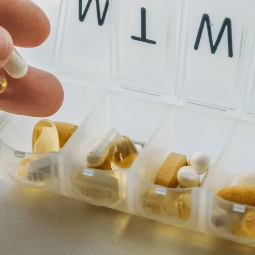 Close-up of a hand reaching for a supplement from a weekly pill organizer filled with various capsules and tablets, implying a daily supplement or medication routine.