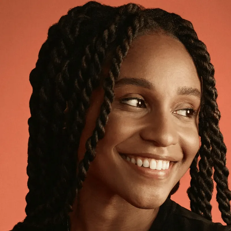 Smiling transgender woman with long braided hair looking to the side against a warm orange background.