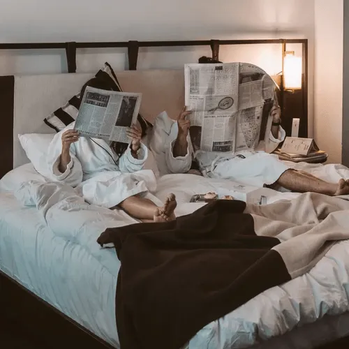 Couple in white bathrobes lying in bed, each holding a newspaper that covers their face, with breakfast on the bed.