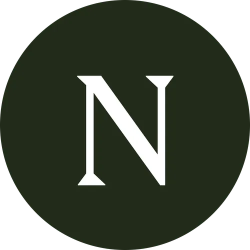 A smiling man and woman pose closely together for a selfie. The man has short curly hair, a beard, and a nose ring, while the woman has long curly hair and is wearing hoop earrings. They are sitting in front of a tiled wall with potted plants in the background. The image is black and white and cropped into a circular frame.