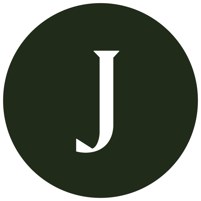 A smiling man and woman pose closely together for a selfie. The man has short curly hair, a beard, and a nose ring, while the woman has long curly hair and is wearing hoop earrings. They are sitting in front of a tiled wall with potted plants in the background. The image is black and white and cropped into a circular frame.