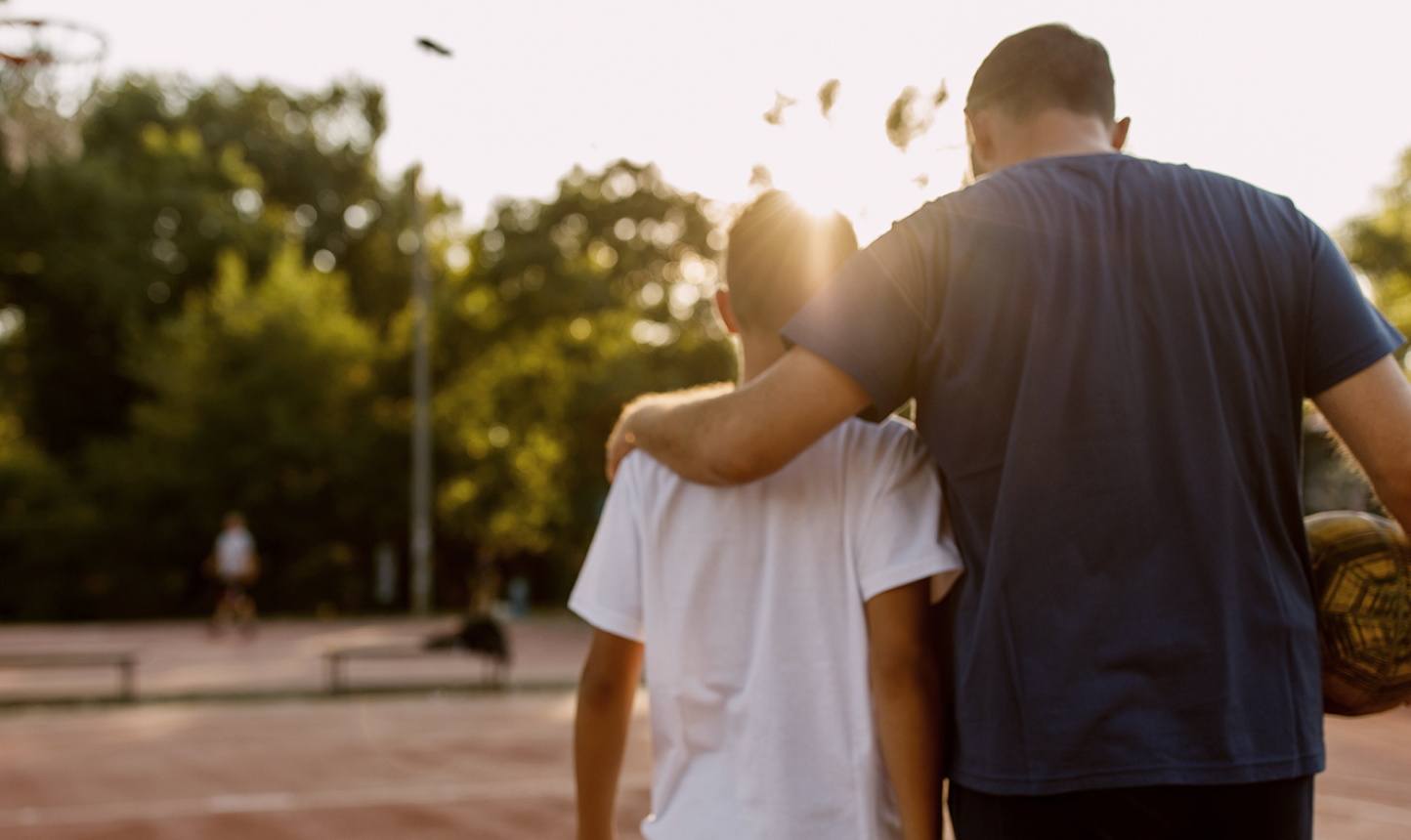 Adult with arm around child holding a soccer ball, standing on a sports court with sunlight filtering through trees.