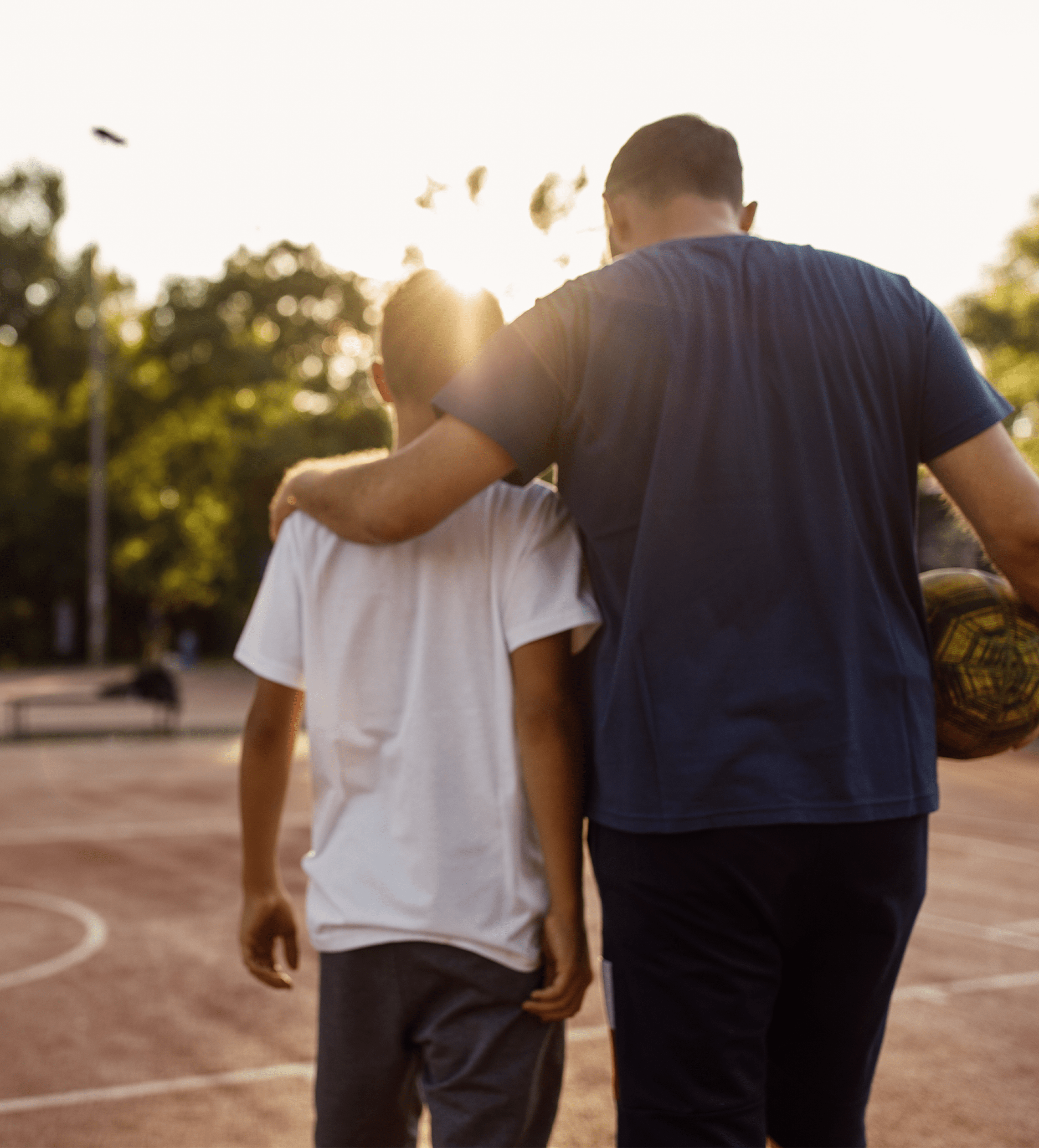 Adult with arm around child's shoulders walking on outdoor basketball court holding a basketball.