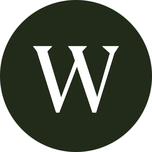 A smiling man and woman pose closely together for a selfie. The man has short curly hair, a beard, and a nose ring, while the woman has long curly hair and is wearing hoop earrings. They are sitting in front of a tiled wall with potted plants in the background. The image is black and white and cropped into a circular frame.