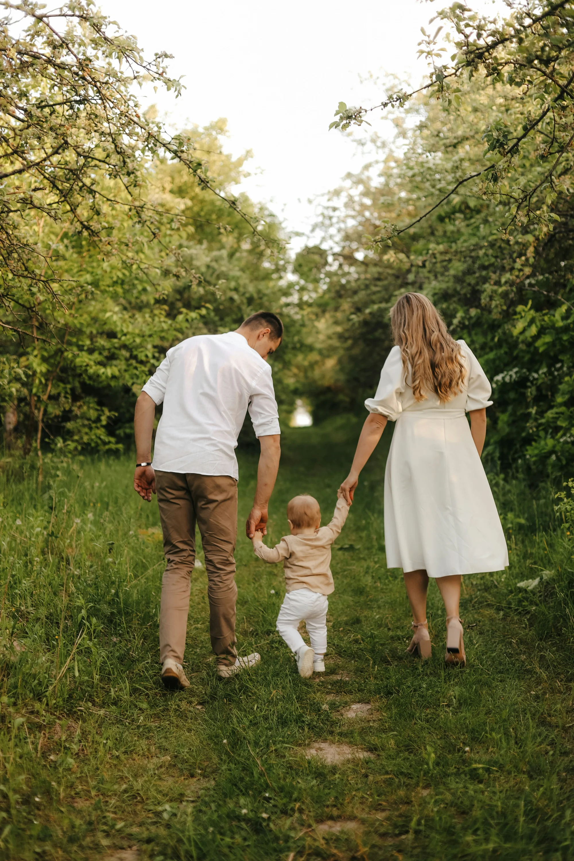 Family of three walking hand-in-hand down tree-lined path, parents in casual wear with toddler between them in natural outdoor setting