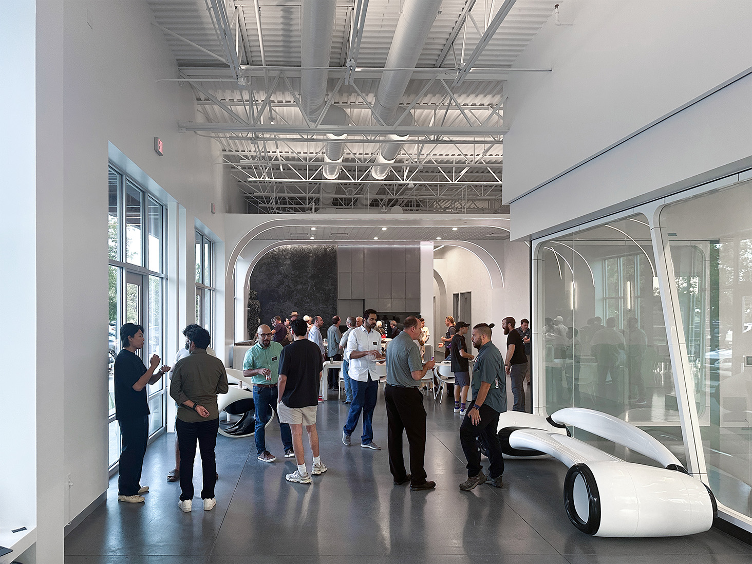 A group of people mingling at an indoor event, engaging in conversation. The room has a modern design with large windows and white arches. 