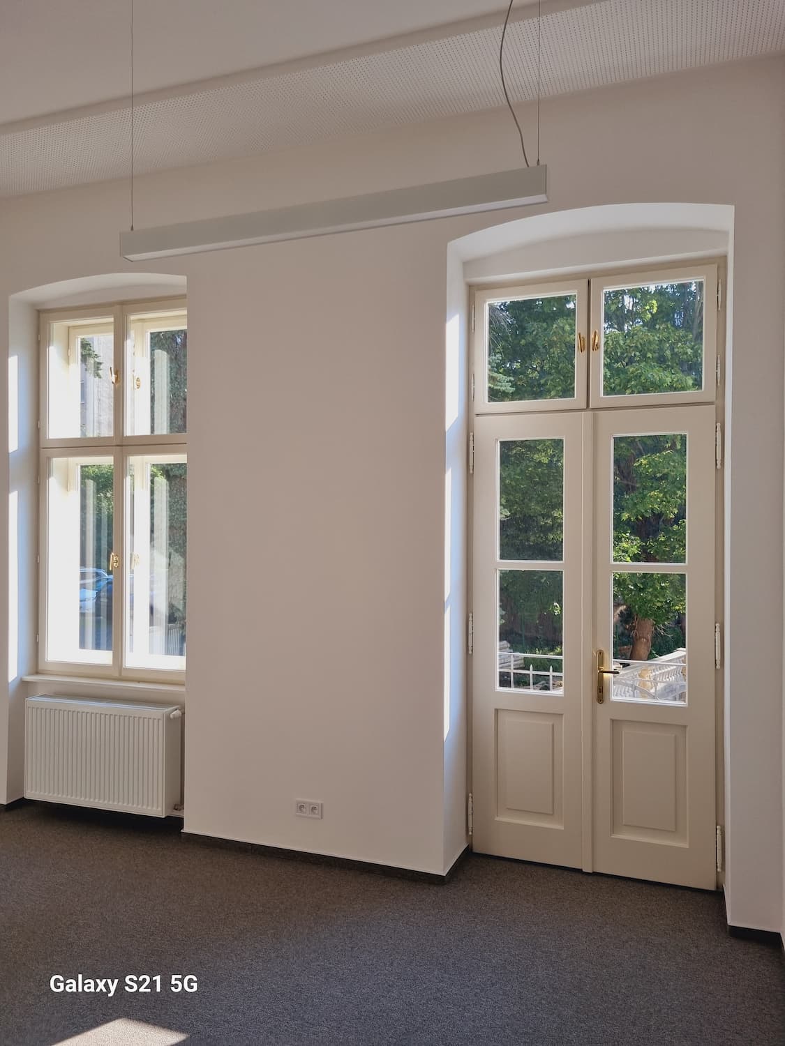 Interiér s vysokým stropom, bielymi stenami, novými rustikálnymi dverami na balkón a moderným radiátorom.