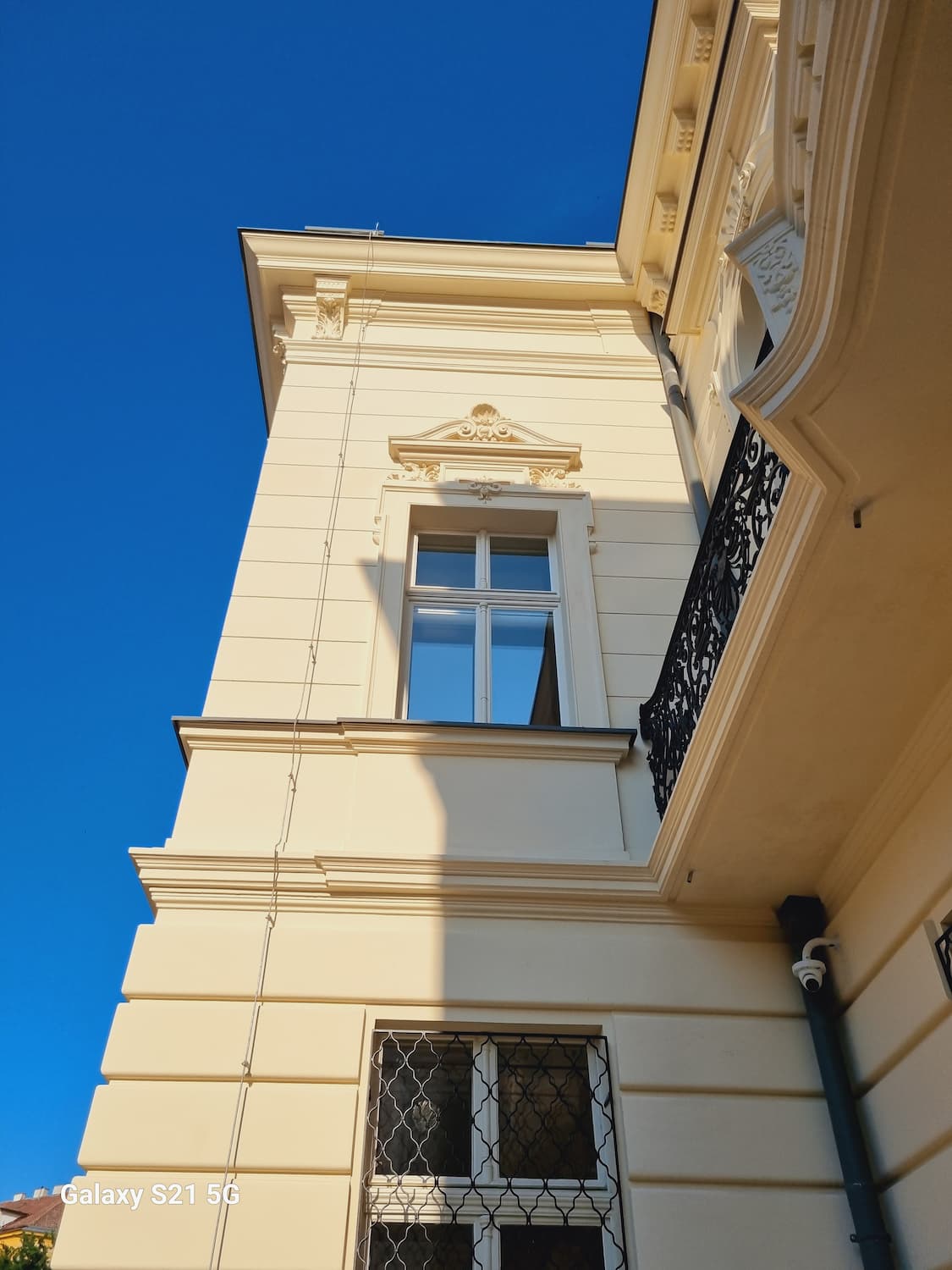 Pohľad na roh budovy, zdôrazňujúci svetlú omietku s kastlovým prepracovaným oknom a modrú oblohu v pozadí.