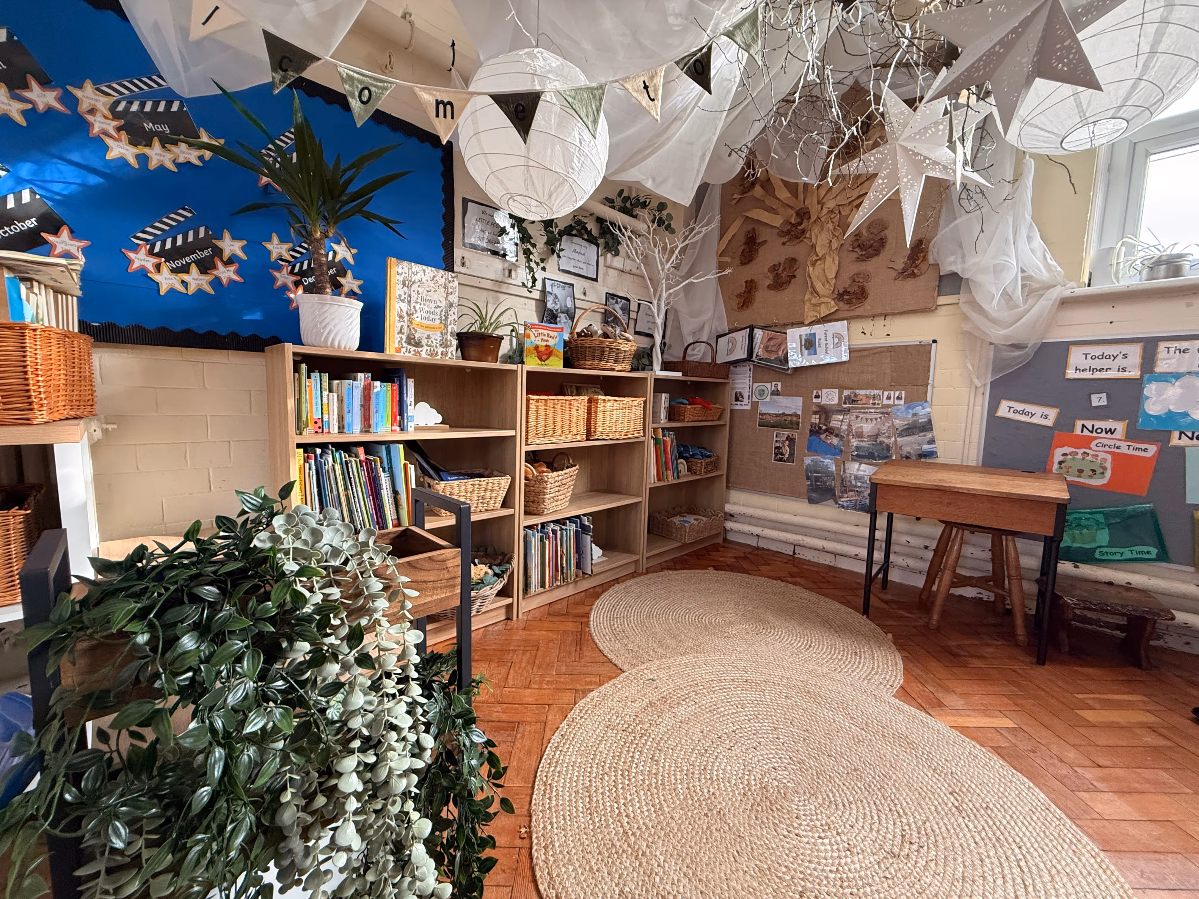 Cozy classroom corner with tall wooden bookshelves filled with books and baskets, a small wooden desk, circular woven rugs on wood floor, plants, and decorative hanging lanterns and stars.