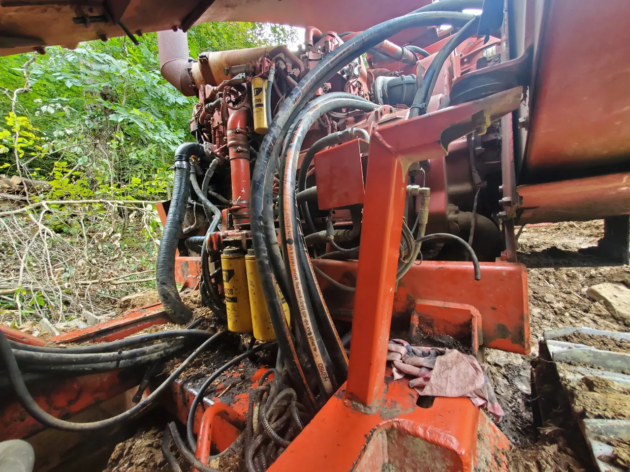 Excavator hydraulic repair with exposed hoses, filters, and engine components on muddy terrain.