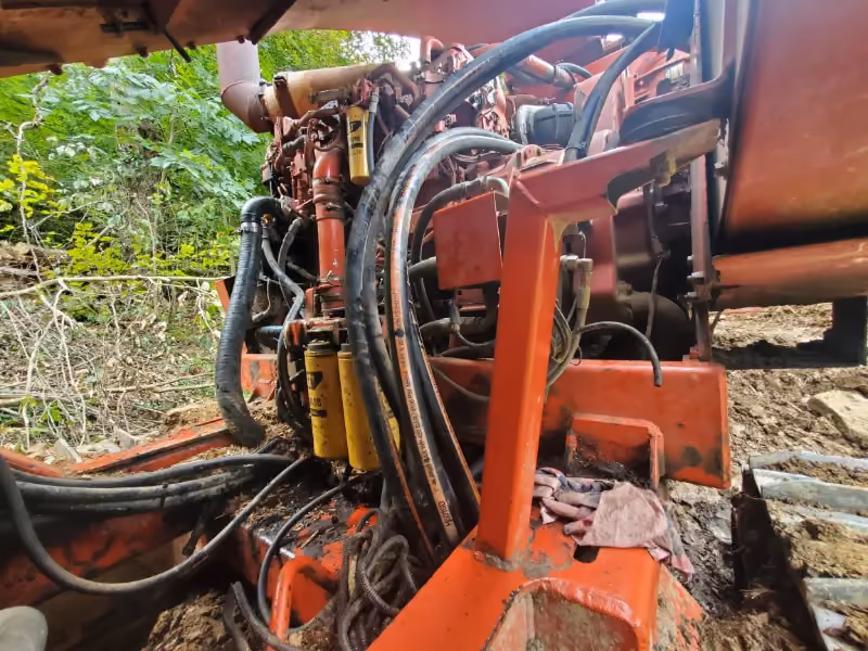 Excavator hydraulic repair with exposed hoses, filters, and engine components on muddy terrain.