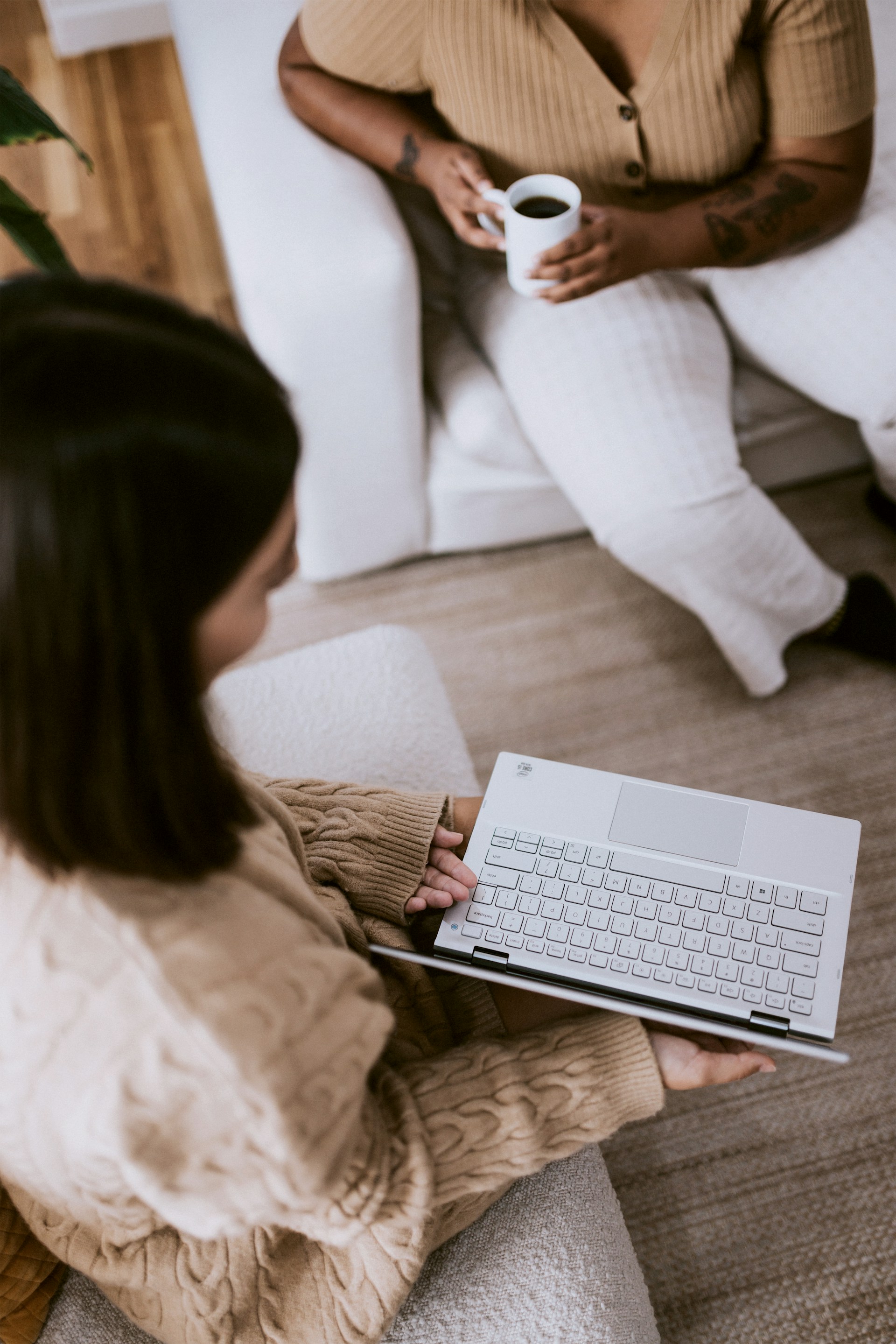 Two people sitting in a cozy setting; one person in a beige sweater holds a white laptop while the other, sitting on a white couch, holds a white mug of coffee.