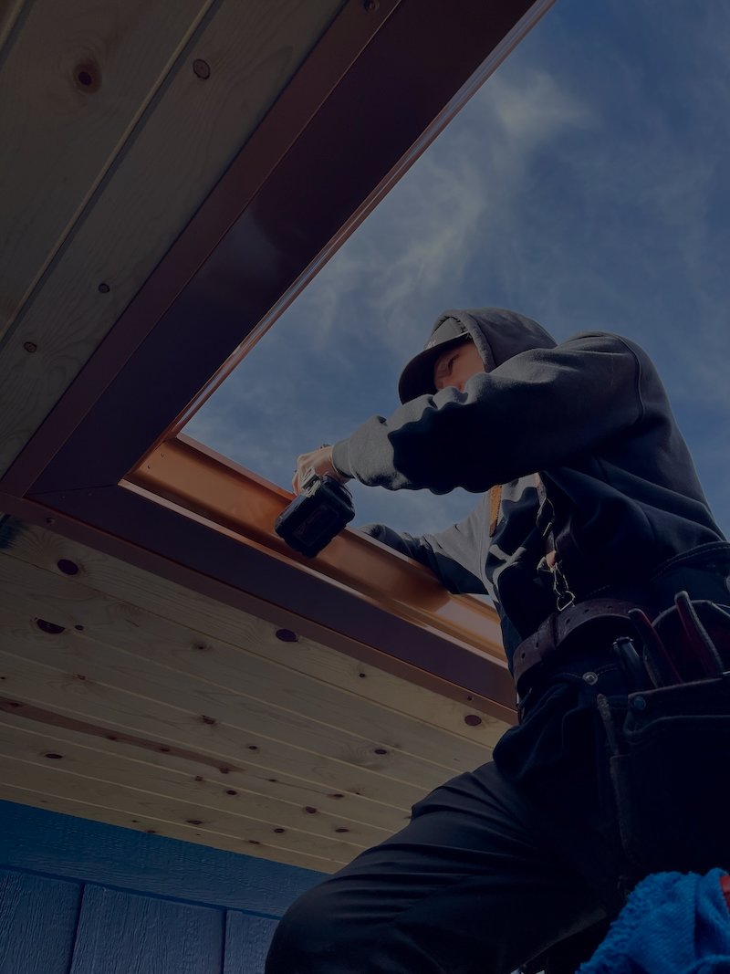 Person wearing a hoodie and cap using a power drill to install a gutter on a wooden roof under a blue sky.