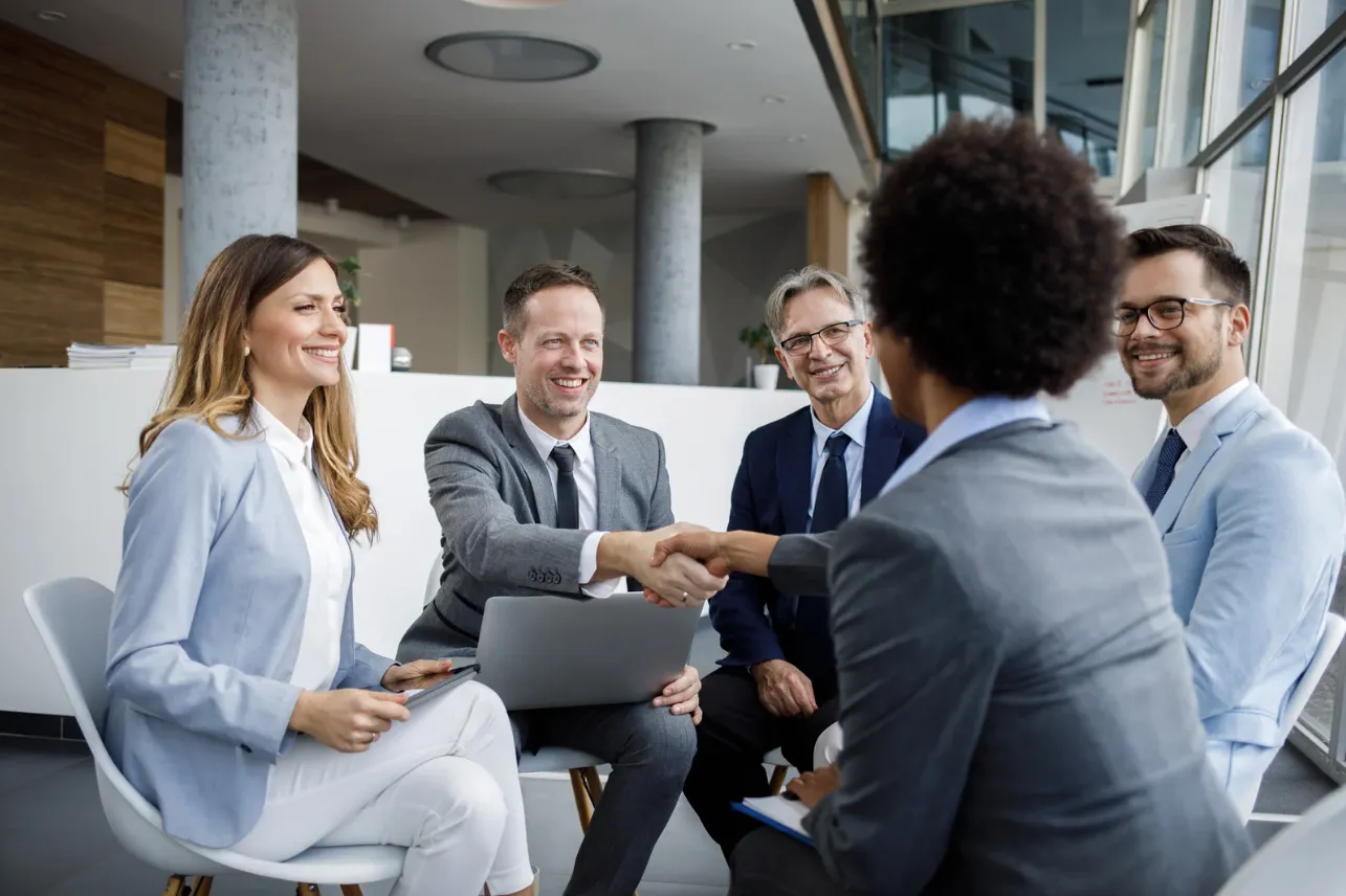 Five business professionals in suits sitting in a modern office, two shaking hands while others smile.