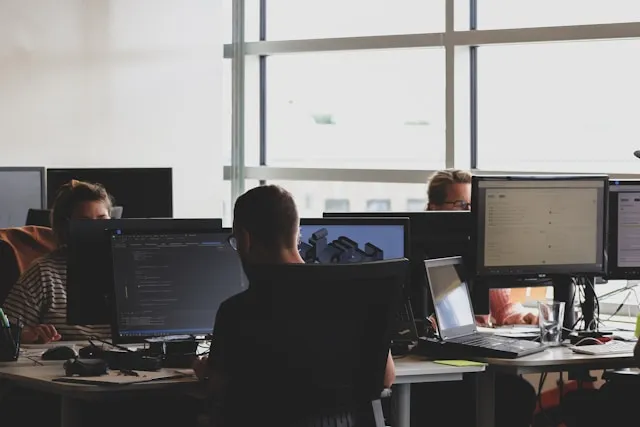 People working at desks with multiple computer monitors in a modern office with large windows.