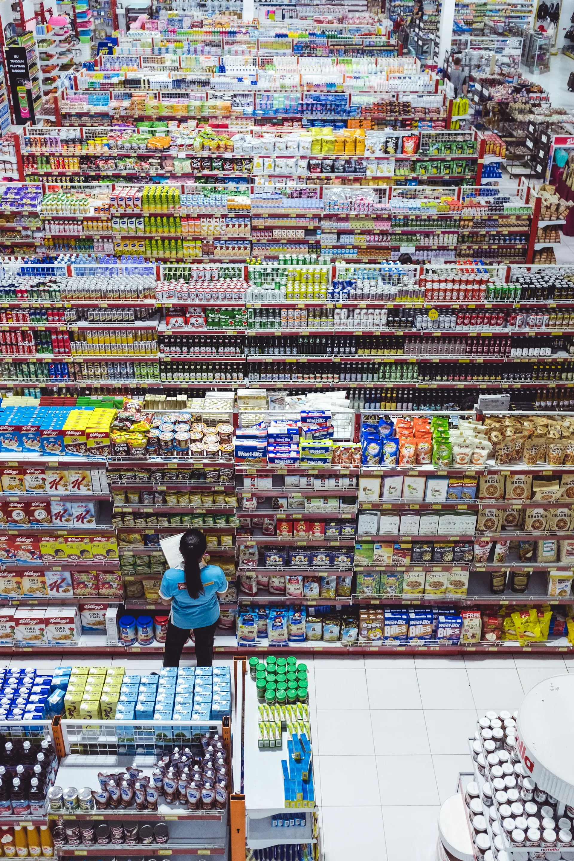 Overhead view of a grocery store aisle filled with various packaged food and drink products, with a person in a blue shirt looking at a product.