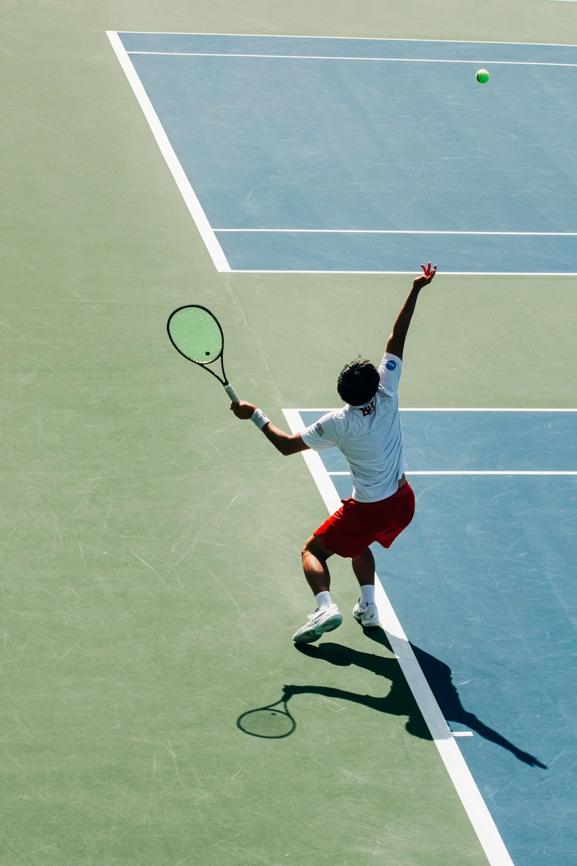 Tennis player in red shorts and white shirt serving a ball on an outdoor hard court.