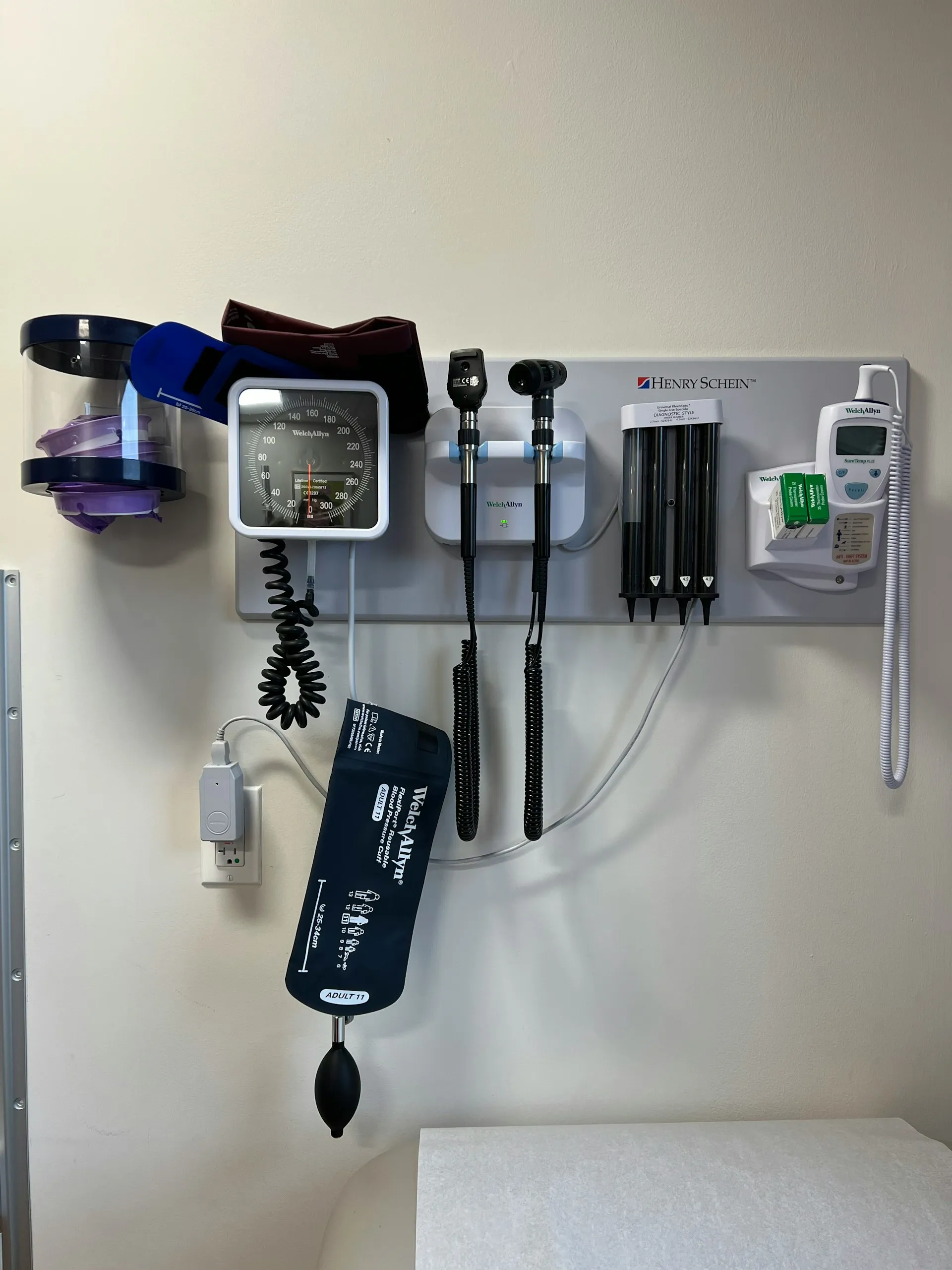 Wall-mounted medical diagnostic equipment including blood pressure cuff, otoscope, ophthalmoscope, thermometer, and disposable specula in a clinic room.