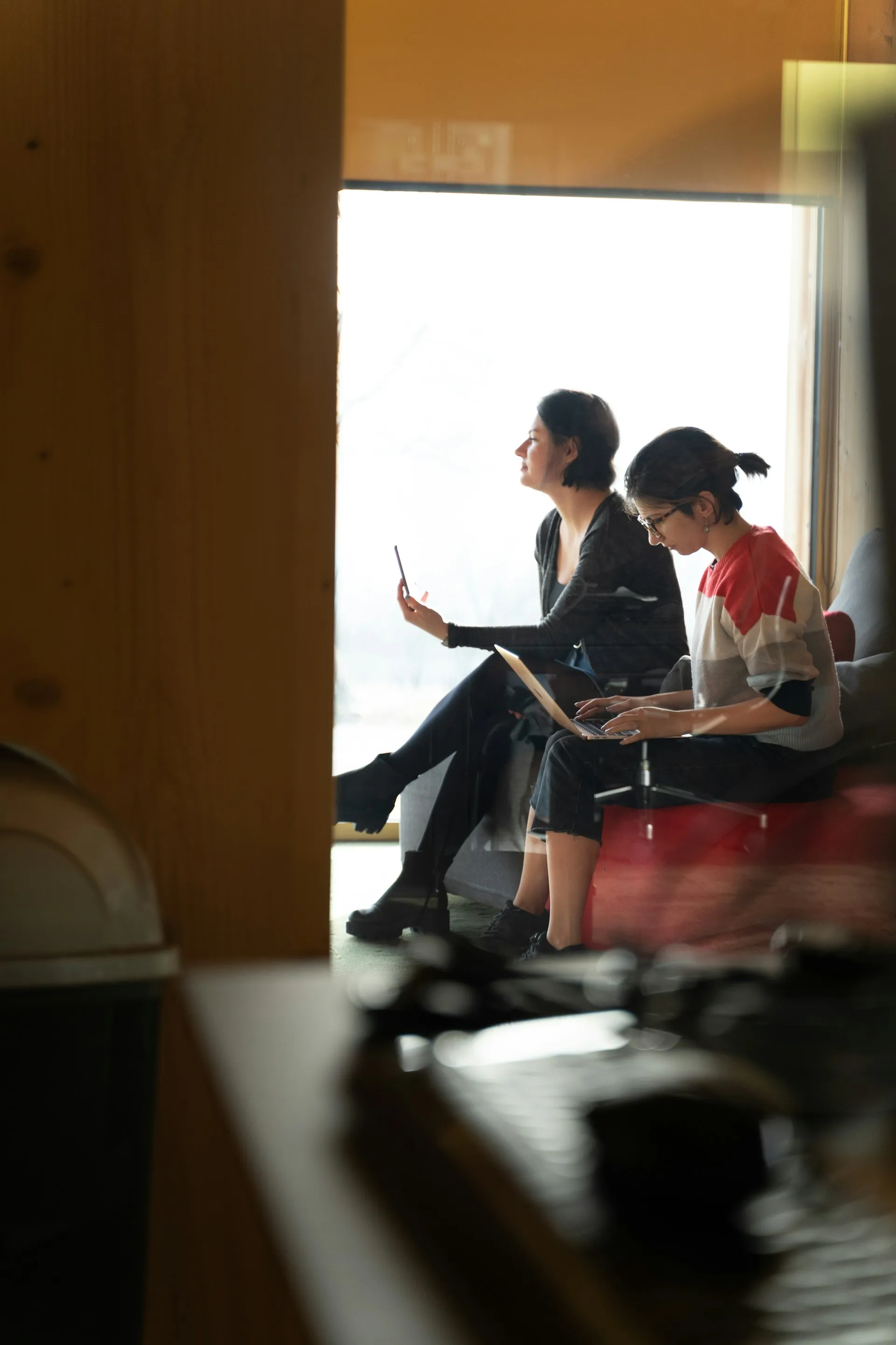 Two women sitting indoors near a window, one using a laptop and the other holding a smartphone.