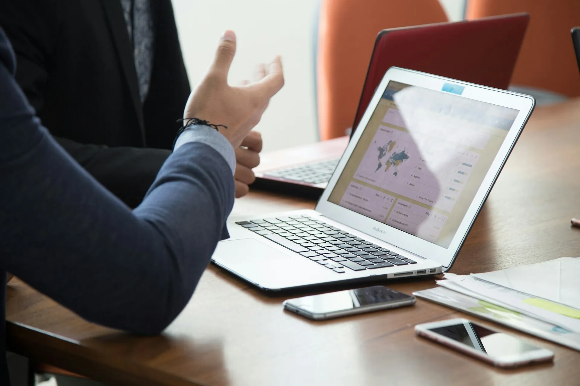Person gesturing with hand while discussing data shown on a MacBook Air screen with a world map and charts on a wooden table.