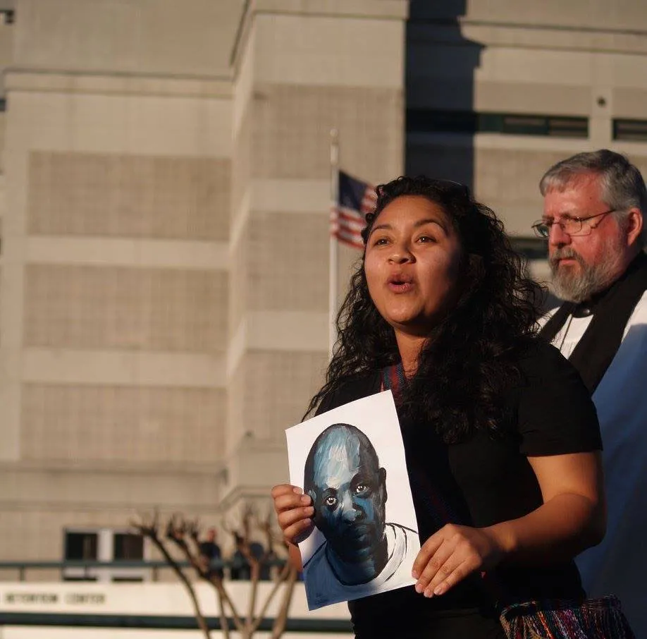 Woman holding portrait of incarcerated man standing in front of Etoway County Detention Center speaking.