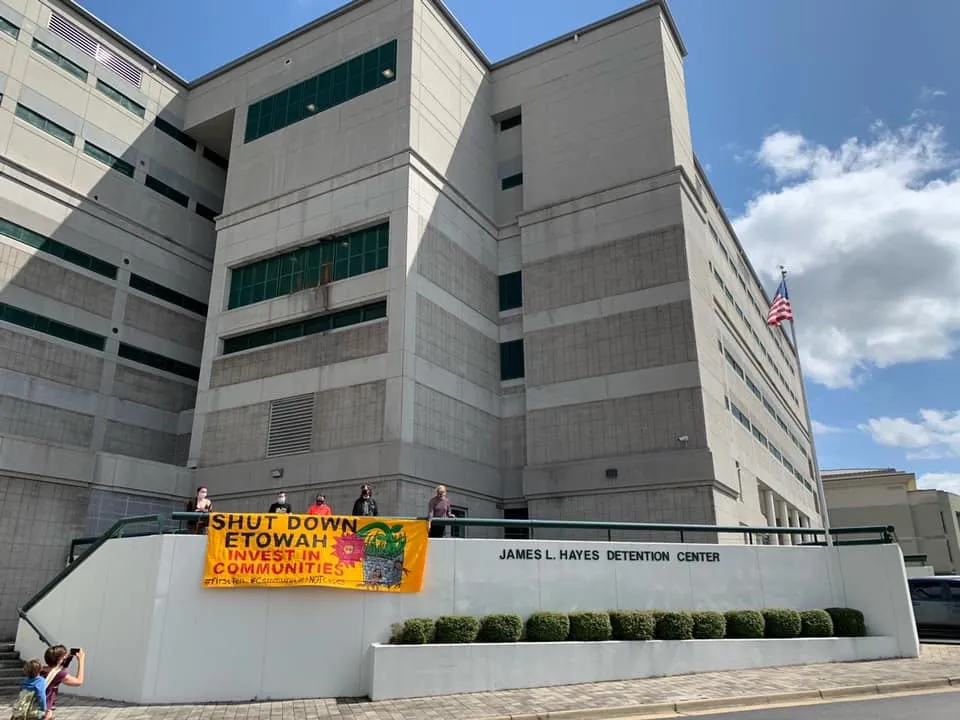 Activists holding banner that reads "Shut Down Etowah. Invest in communities" in front of Etowah County Detention Center.