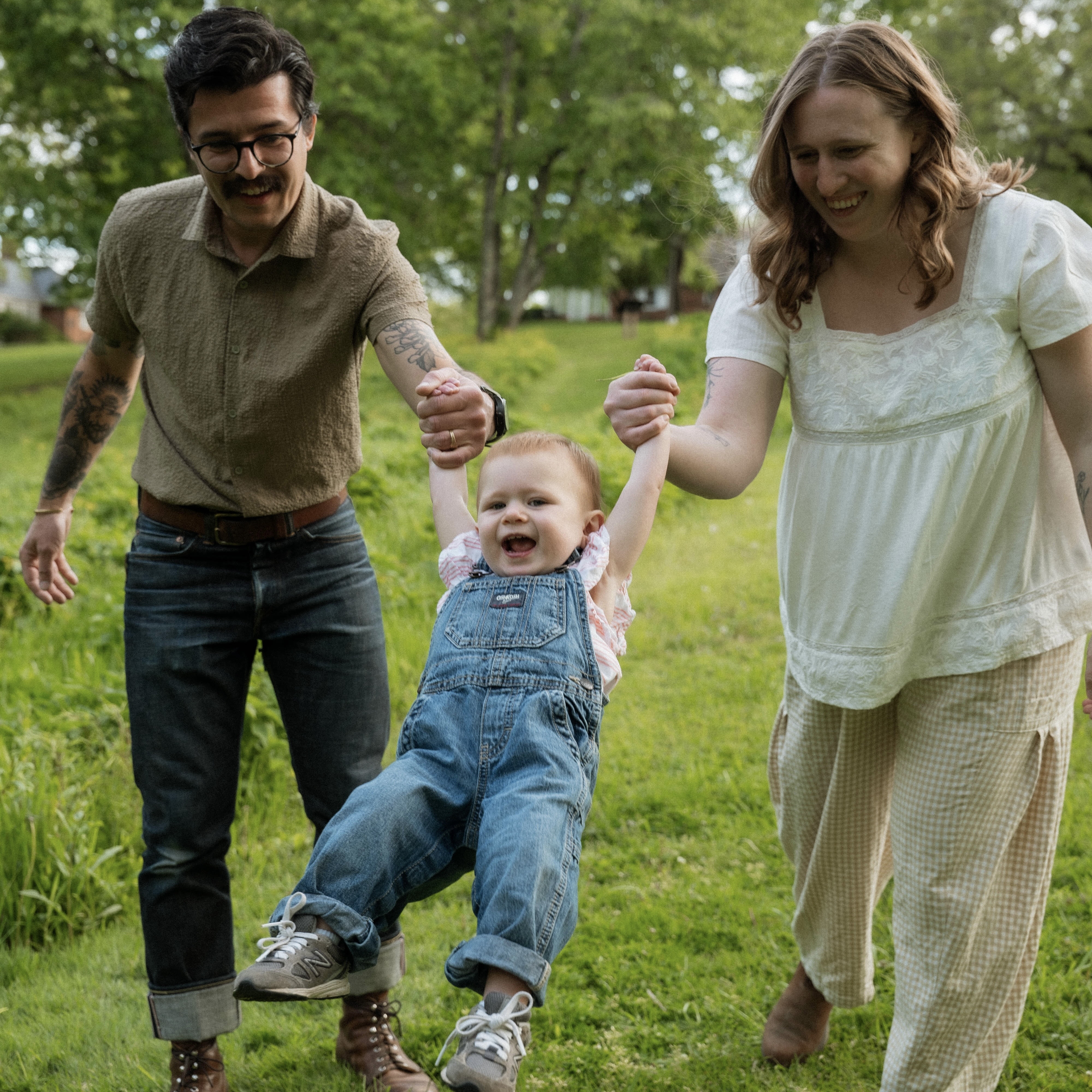 Happy baby swinging by holding hands of smiling adults walking on grass in a park.