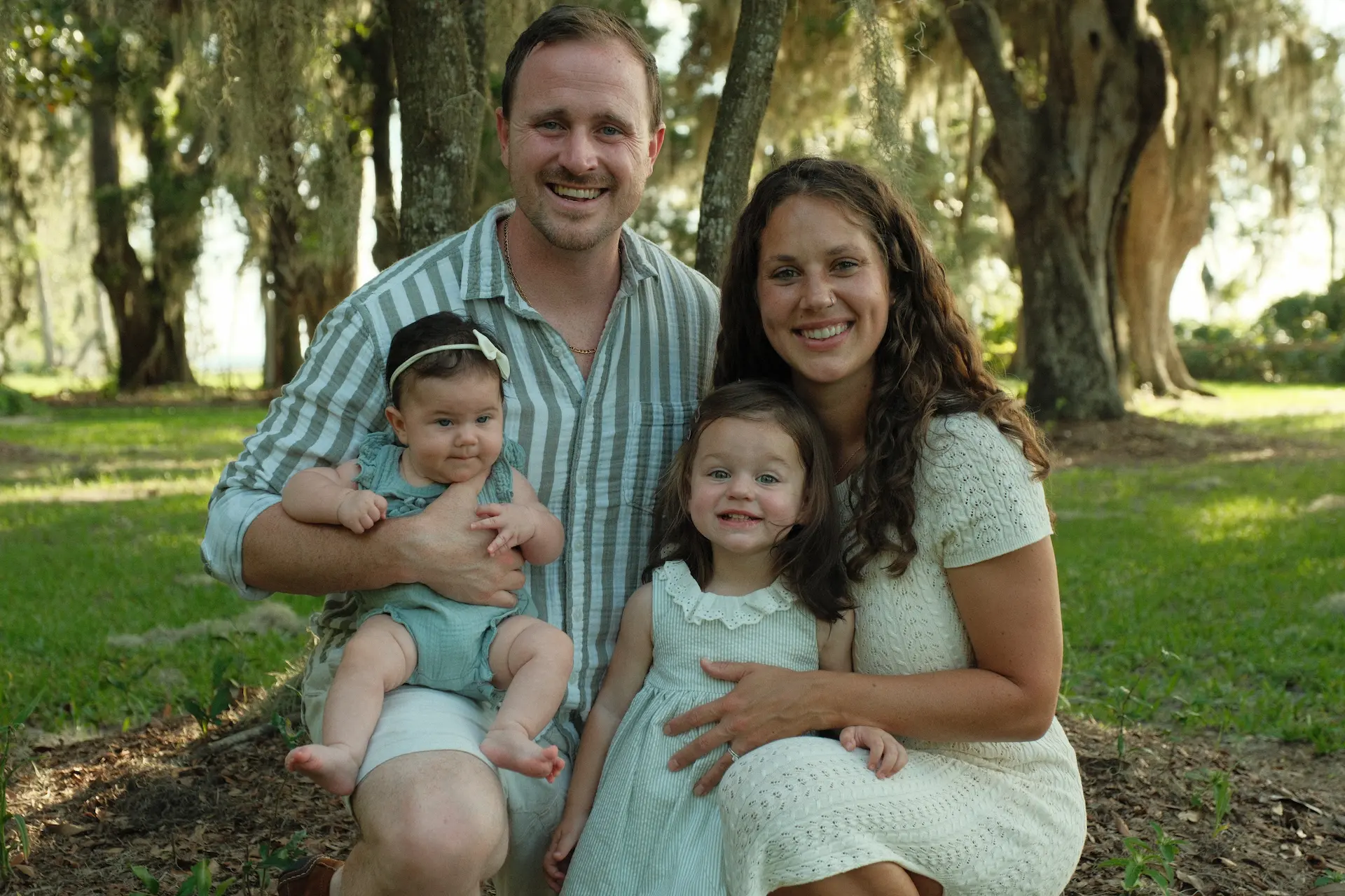 Smiling family of four outdoors with a man holding a baby girl wearing a blue outfit and headband, and a woman kneeling beside a toddler girl in a white dress, with trees and green grass in the background.
