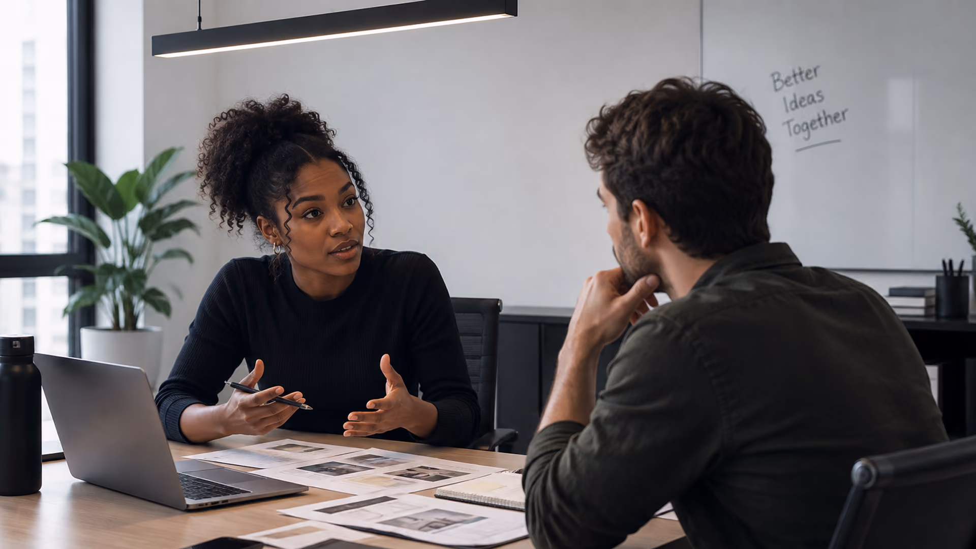 Two professionals discuss documents over a laptop in an office with a whiteboard reading 'Better Ideas Together'.