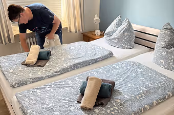 Person richtet ein Bett mit grauen gemusterten Bettdecken und zusammengerollten Handtüchern.