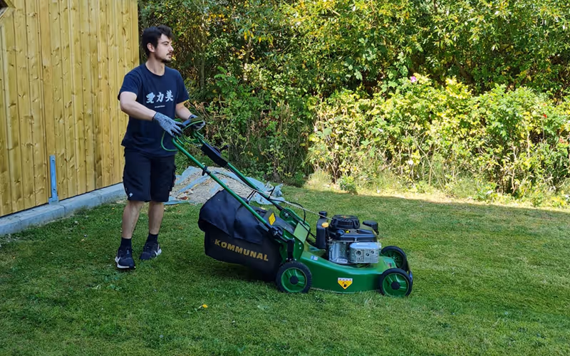 Mann mäht Rasen mit grünem Benzin-Rasenmäher neben Holzwand und grünen Büschen.