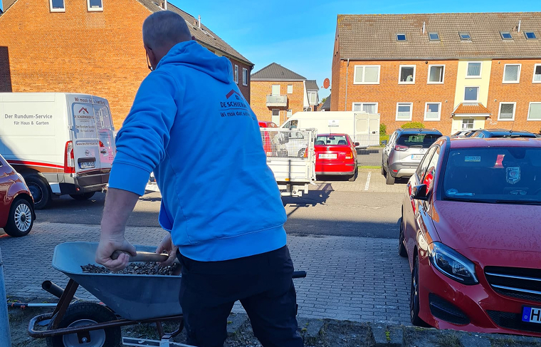 Mann in blauer Kapuzenjacke schiebt Schubkarre mit Schotter in einem Wohngebiet mit geparkten Autos und Häusern im Hintergrund.