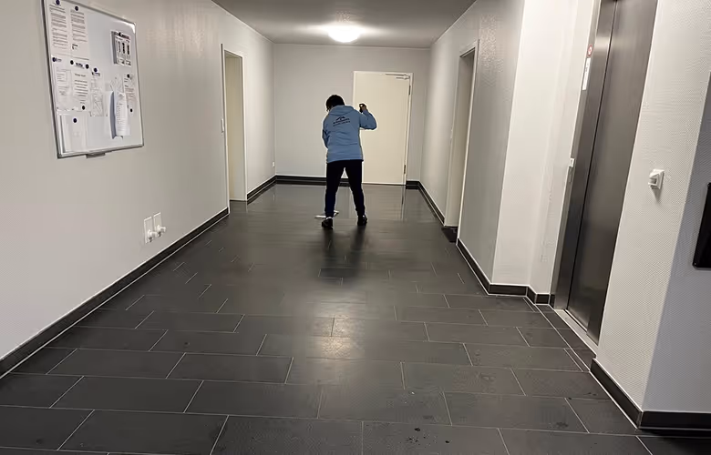 Person reinigt den Boden in einem hellen Flur mit dunkelgrauen Fliesen und weiß gestrichenen Wänden.