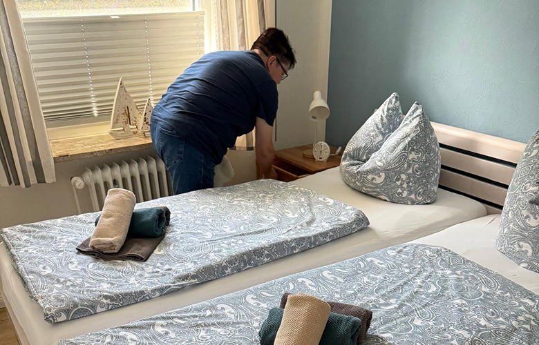 Person richtet Bett in einem hellen Schlafzimmer mit zwei Betten und gemusterten Bettbezügen her.