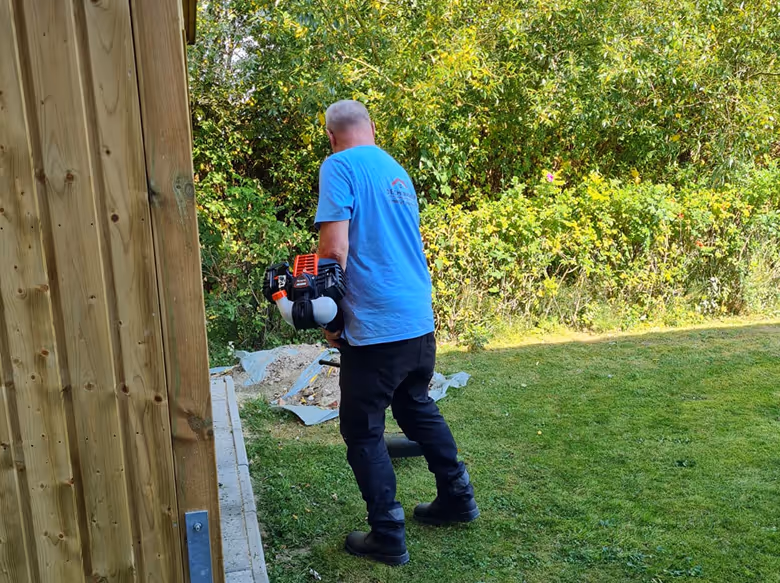 Mann mit blauem T-Shirt und schwarzen Hosen bedient einen Laubbläser im Garten neben einem Holzhaus.