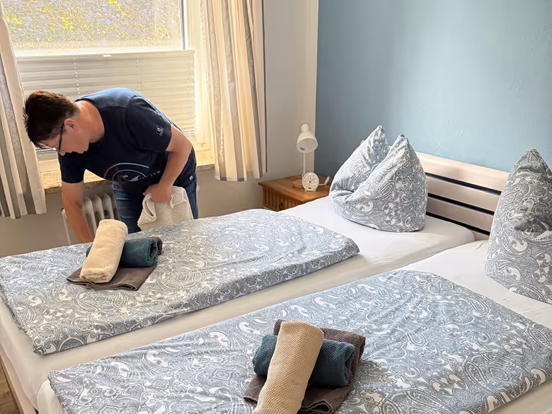 Eine Person richtet zwei Betten mit gemusterten Bettdecken und zusammengerollten Handtüchern in einem hellen Schlafzimmer mit blau gestrichener Wand her.