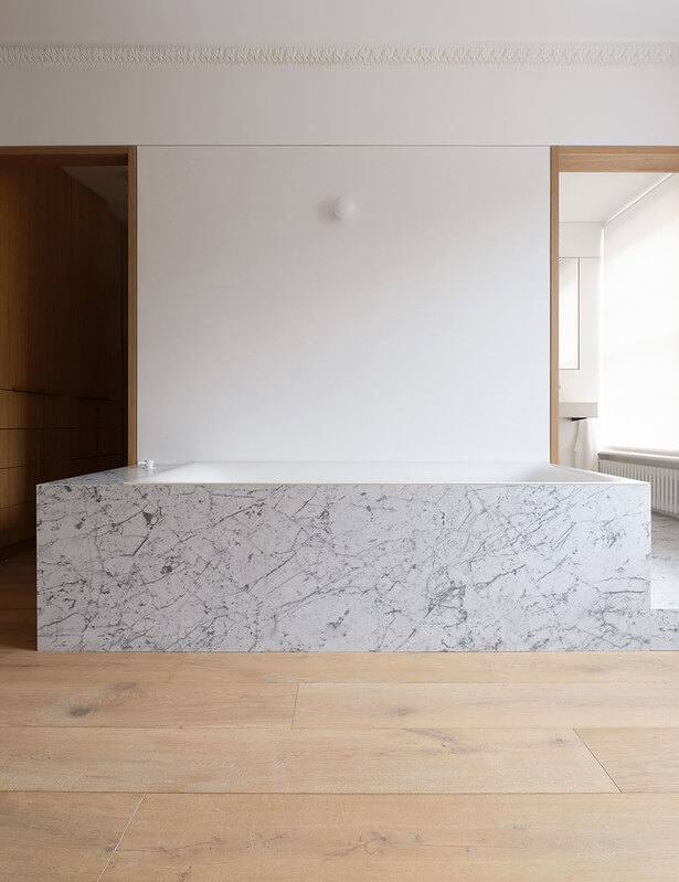 Modern bathroom with a large rectangular marble bathtub, wooden floor, and minimalist white wall with a small round light fixture.