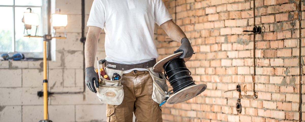 A man wearing a white shirt and khaki  work pants with a full tool belt walks through a brick warehouse carrying a large roll of black electrical wire.