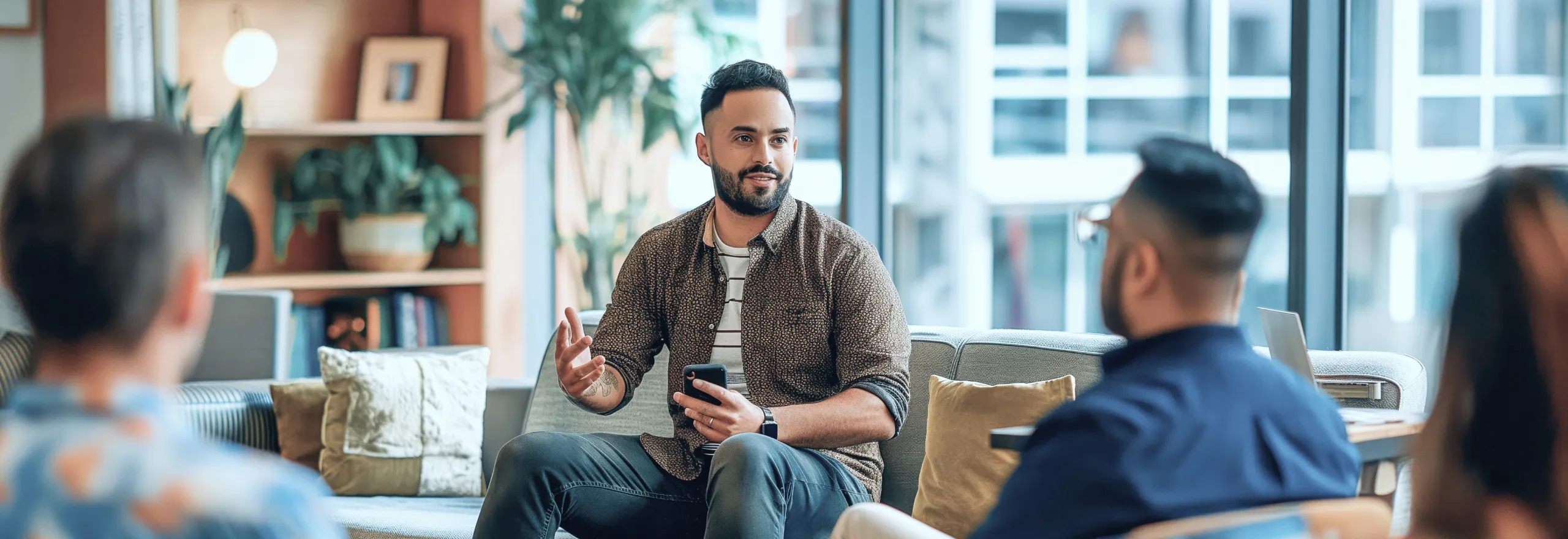 Man speaking while holding a phone during a casual group discussion in a modern office lounge.