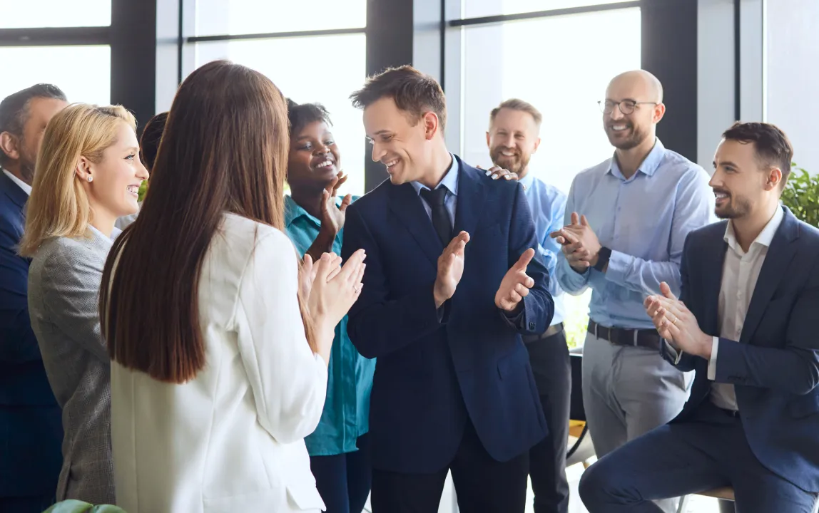 A group of business professionals standing together in an office, smiling and applauding a colleague in a suit.