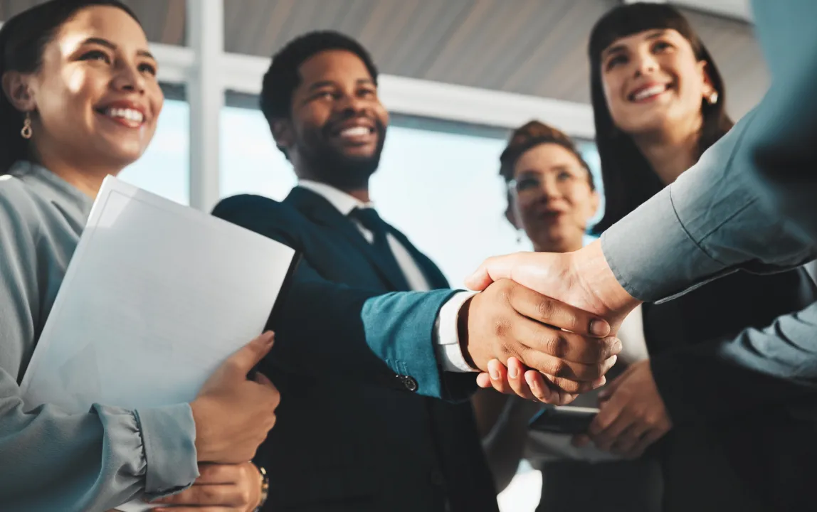 Business professionals smiling while two people shake hands in agreement during a meeting.