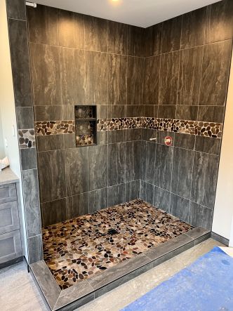 A walk in shower with a colourful pebble floor, a dark large format tile on the walls and a pebble accent band through the middle of the shower that matches the floor.