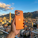 Hand holding an orange iPhone over a cityscape with mountains in the background during sunset.