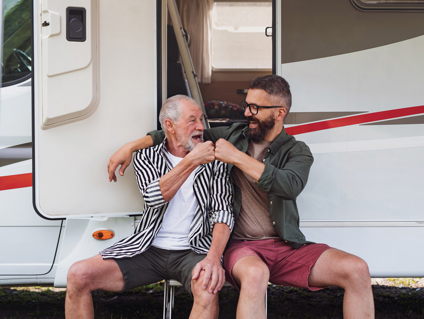 Father and son sitting outside a caravan, fist bumping and laughing together, an image featured in the CIL Weekends campaign celebrating family bonds.
