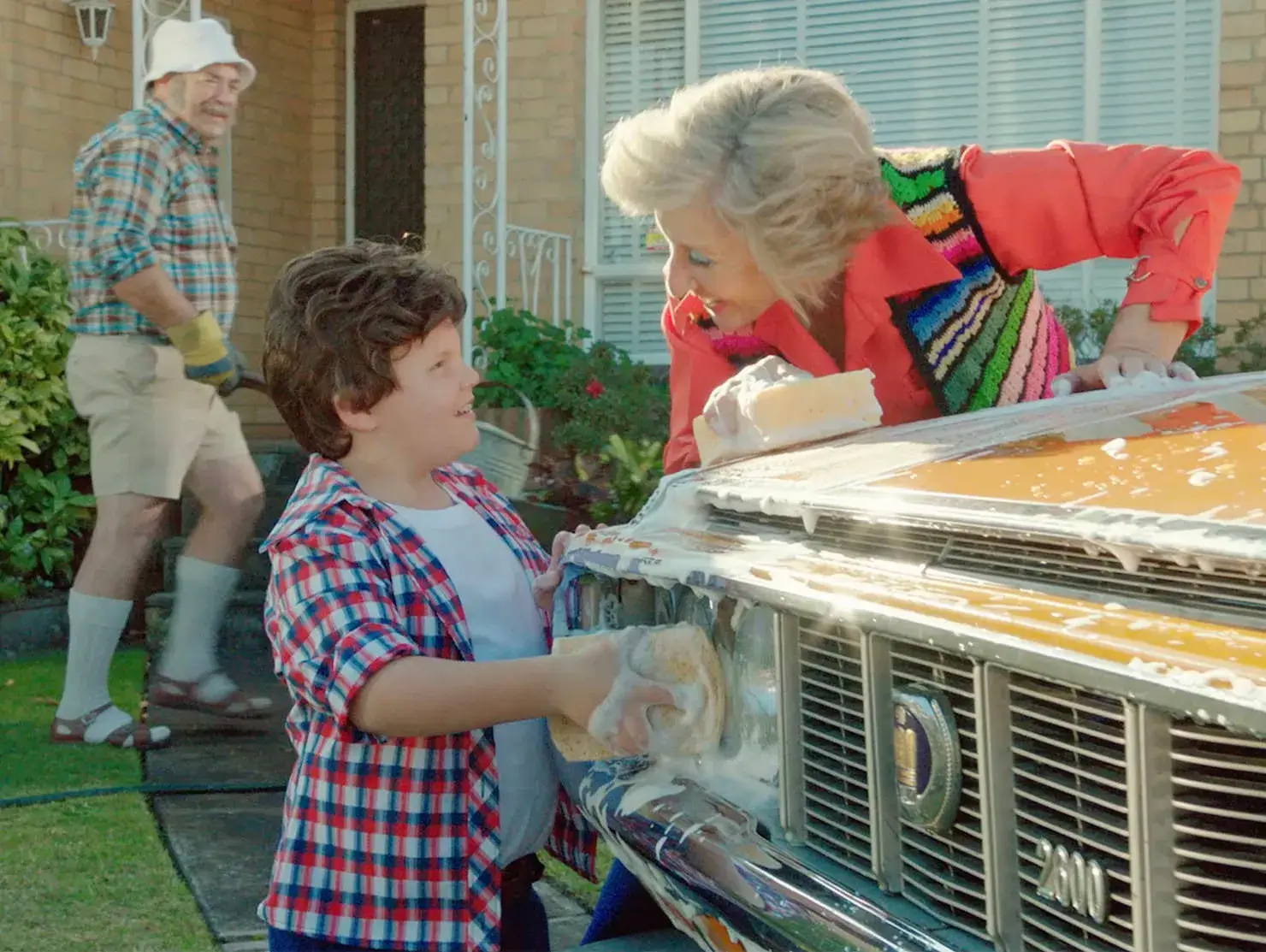 Young boy and grandmother washing a Toyota Crown together, with grandfather gardening in the background, scene from Shannons Insurance for Motoring Enthusiasts TV ad.