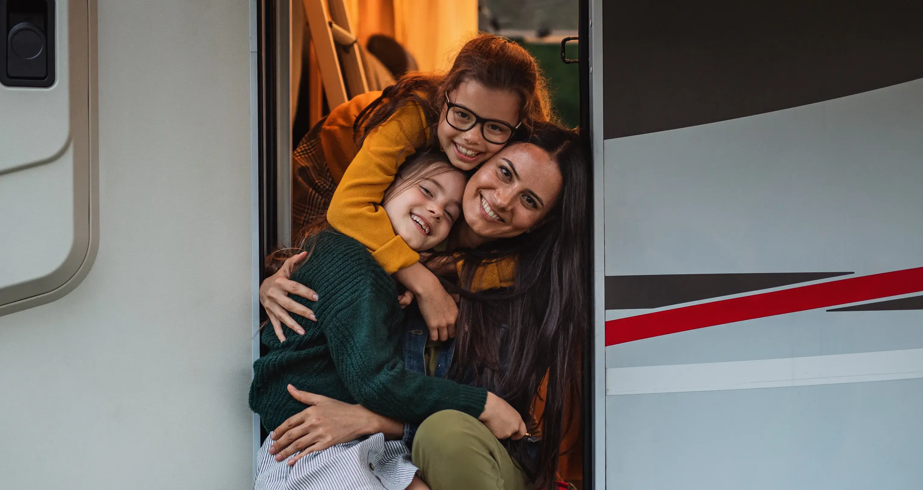 Mother and two daughters smiling and hugging in the doorway of a caravan during a holiday escape, part of the CIL Weekends campaign celebrating memorable getaways.