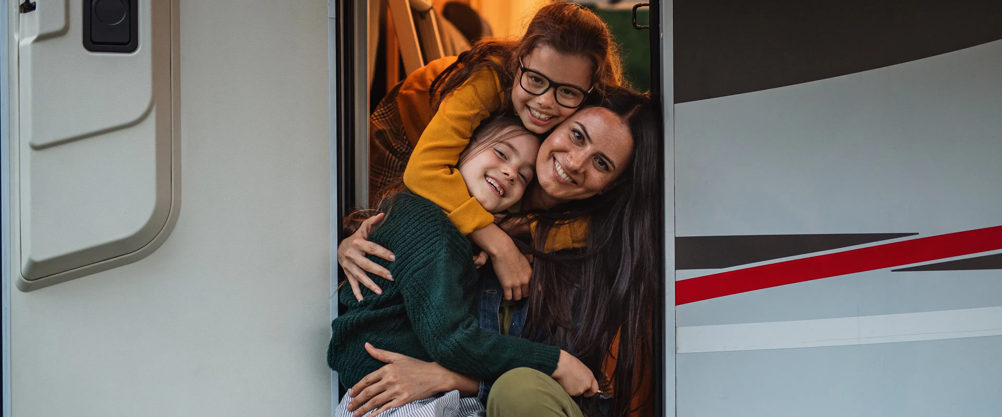 Mother and two daughters smiling and hugging in the doorway of a caravan during a holiday escape, part of the CIL Weekends campaign celebrating memorable getaways.
