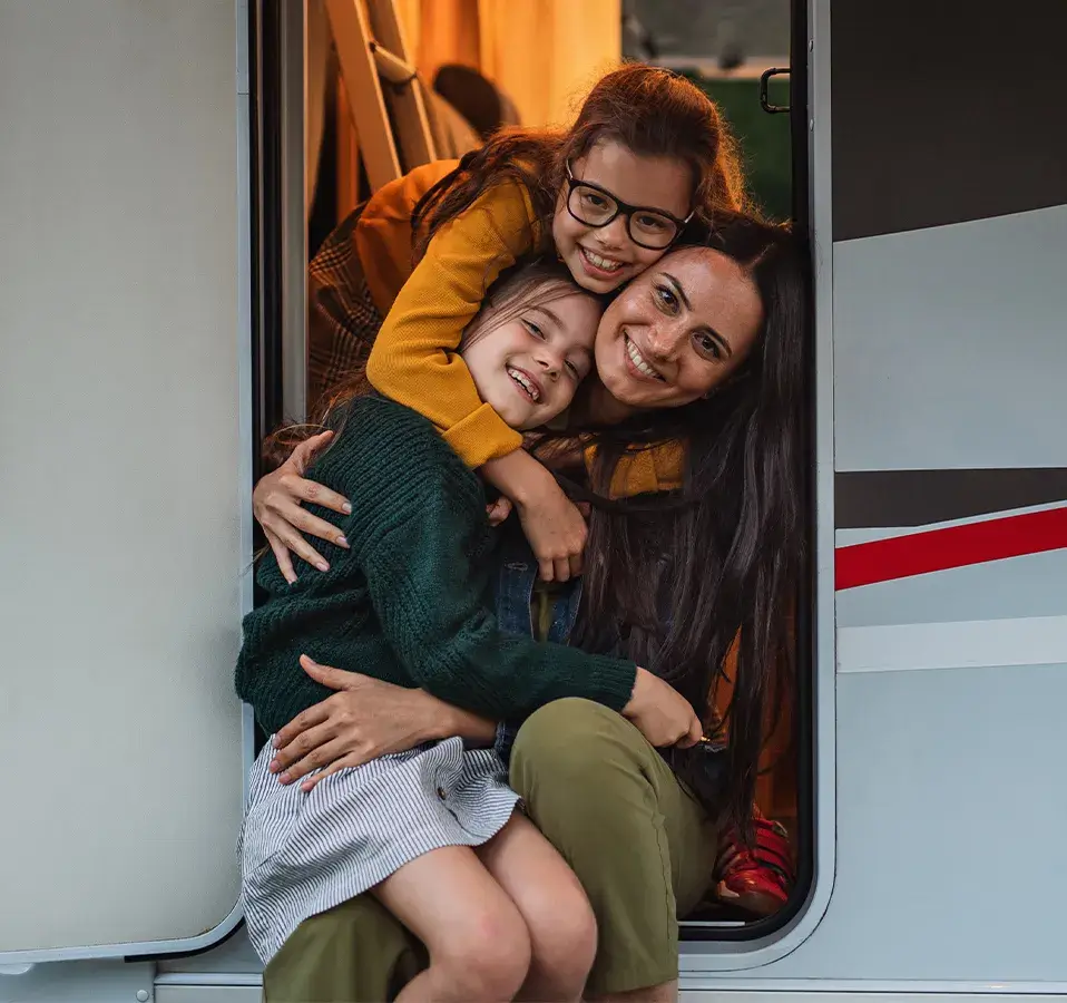 Mother and two daughters smiling and hugging in the doorway of a caravan during a holiday escape, part of the CIL Weekends campaign celebrating memorable getaways.