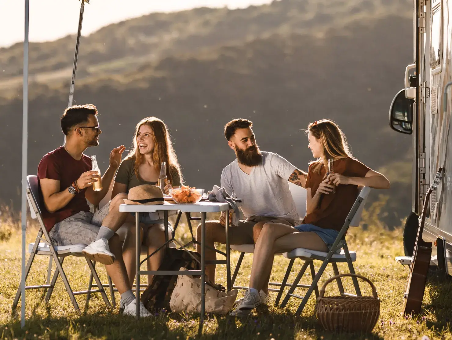 Group of young friends sharing drinks and laughter around a camping table beside their caravan, captured as part of the CIL Weekends campaign encouraging outdoor adventures.